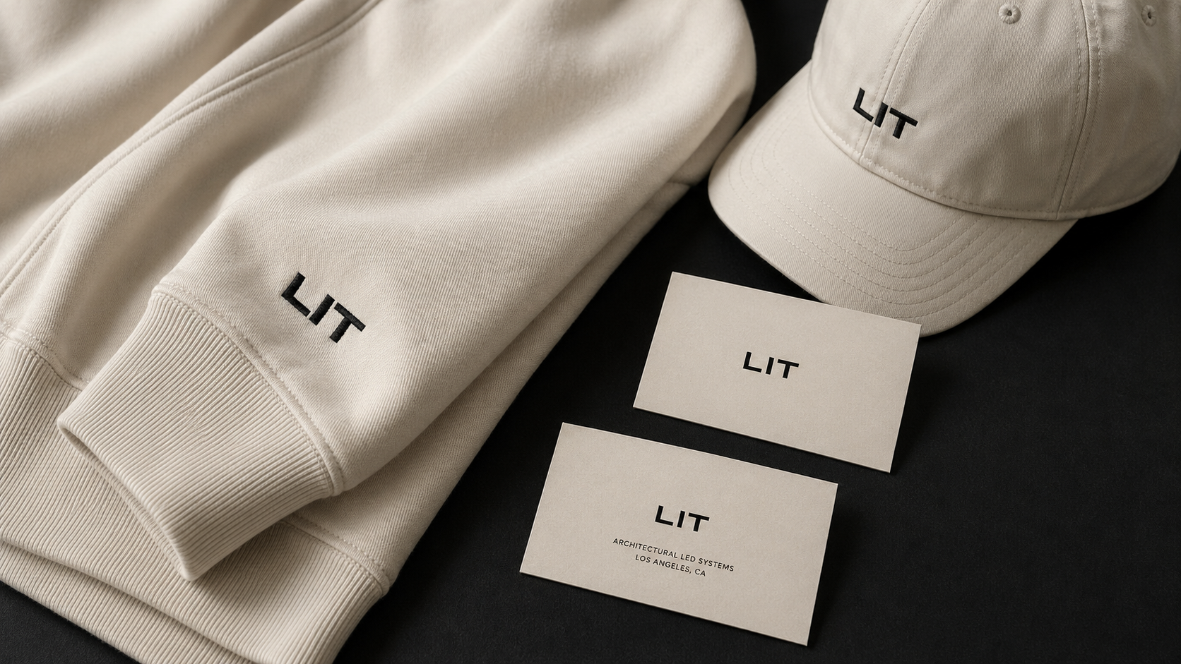 Detail of a cream Lit Group hoodie sleeve with embroidered LIT mark, paired with a cream cap and stationery.